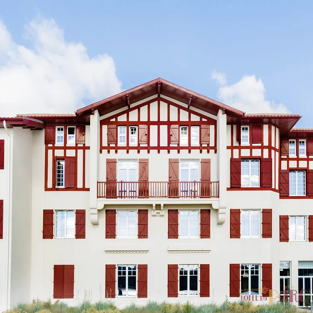 Vue frontale de la façade de l’Hôtel du Parc Hossegor, bâtiment de style basco-landais avec colombages rouges et volets assortis, sous un ciel bleu dégagé.